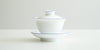 Side view of a white porcelain gaiwan, with blue rings painted around the edges of the plate, bowl, and the handle of the lid. The sides are rounded at the bottom before curving out towards the lip.
