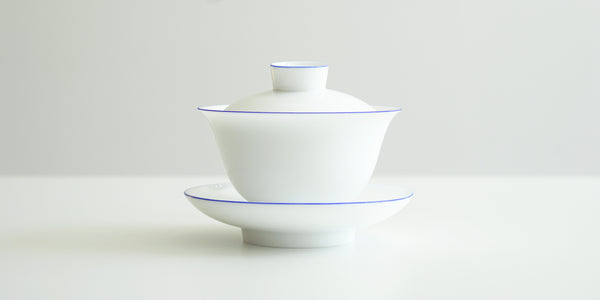Side view of a white porcelain gaiwan, with blue rings painted around the edges of the plate, bowl, and the handle of the lid. The sides are rounded at the bottom before curving out towards the lip.