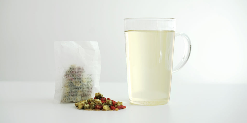 A sachet containing chrysanthemum buds, goji berries, and winter melon sugar. A pile of the same ingredients on the white table. A glass cup filled with the brewed tea.