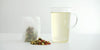 A sachet containing chrysanthemum buds, goji berries, and winter melon sugar. A pile of the same ingredients on the white table. A glass cup filled with the brewed tea.