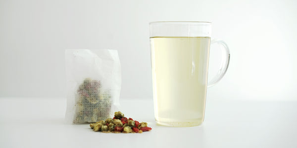 A sachet containing chrysanthemum buds, goji berries, and winter melon sugar. A pile of the same ingredients on the white table. A glass cup filled with the brewed tea.