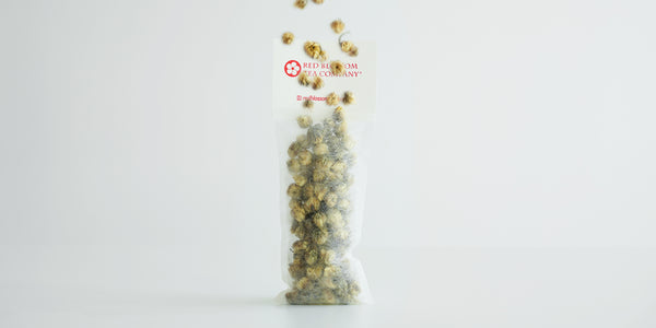 A tea sachet standing filled with chrysanthemum buds. More chrysanthemum buds are raining down from above.