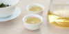 Two small teacups of white tea, with a gaiwan in the background on the left and a glass server on the right.