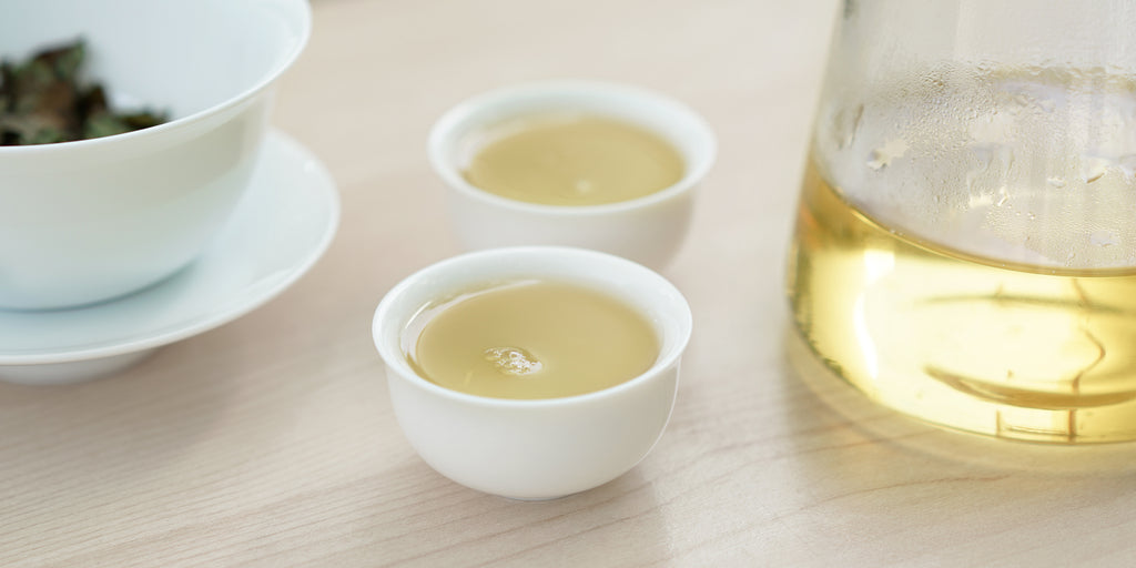 Two small teacups of white tea, with a gaiwan in the background on the left and a glass server on the right.
