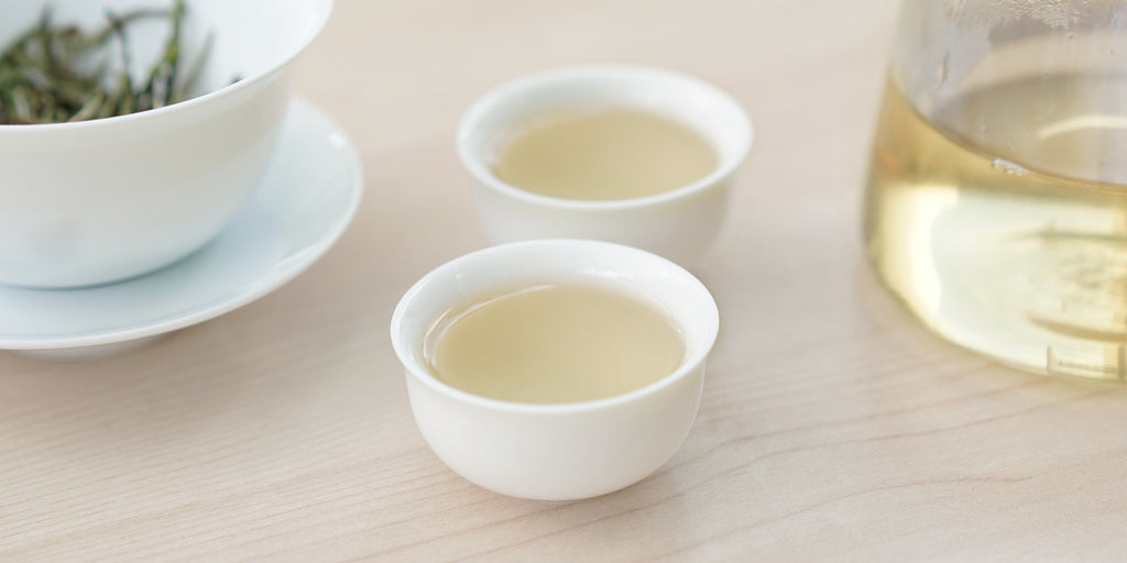Two small teacups of white tea, with a gaiwan in the background on the left and a glass server on the right.