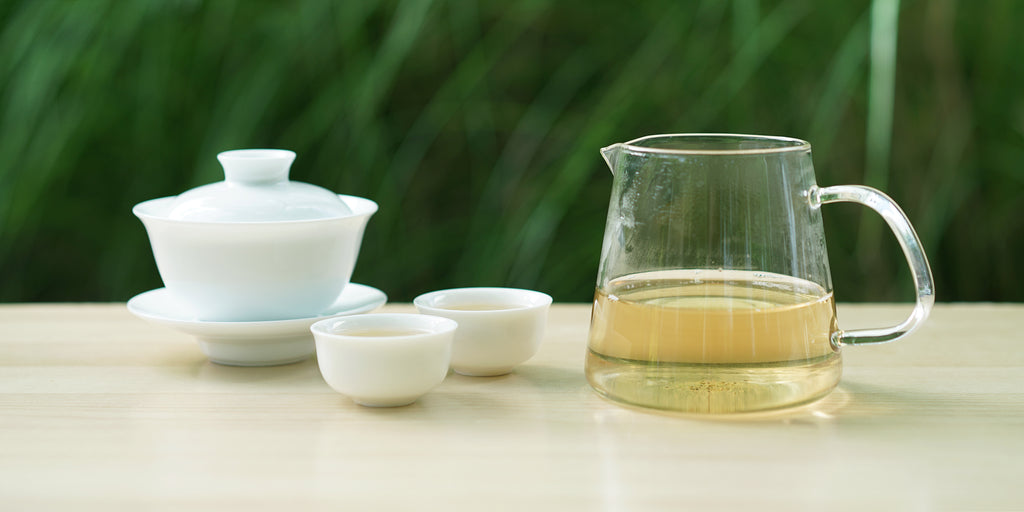 From left to right, a gaiwan, two teacups, and a glass server. The teacups and server have white tea in them.