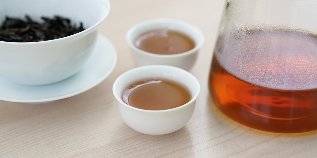 Two small teacups of Wuyi oolong tea, with a gaiwan in the background on the left and a glass server on the right.