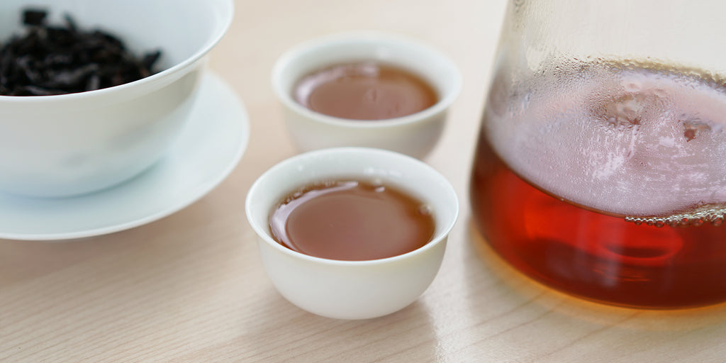 Two small teacups of Wuyi oolong tea, with a gaiwan in the background on the left and a glass server on the right.