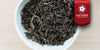 A white dish containing dry pu-erh tea leaves. A red ribbon in the top right corner reads “Red Label Collection”