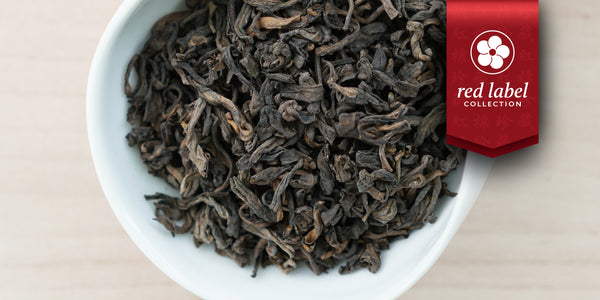 A white dish containing dry pu-erh tea leaves. A red ribbon in the top right corner reads “Red Label Collection”