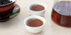 Two small teacups of pu-erh tea, with a gaiwan in the background on the left and a glass server on the right.